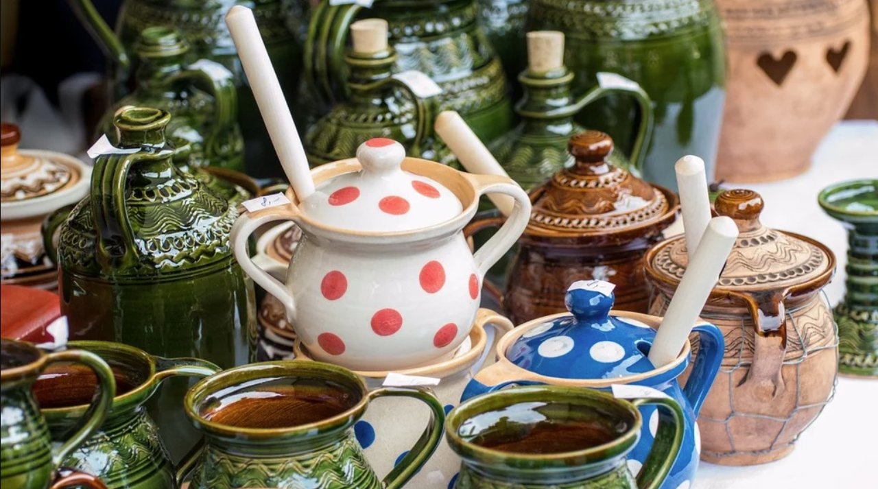 A still life of slip-glazed earthenware — green-and-amber jugs, polka-dotted lidded pots and rustic mugs gathered on a table.